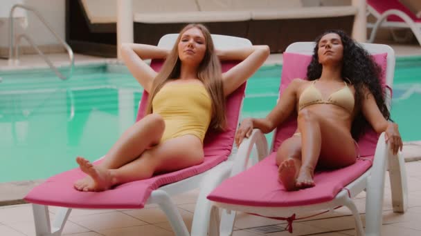 Привлекательная молодая многонациональная латиноамериканка-кавказка подруги туристки модели в купальниках расслабляются вместе загорая на шезлонге возле бассейна спа-отеля наслаждаясь летним роскошным отдыхом - Кадры, видео