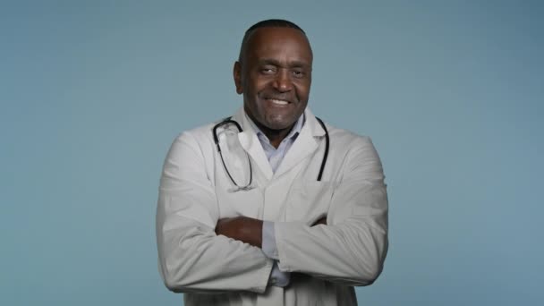 Ältere selbstbewusste glückliche Arzt steht in grauen Hintergrund Studio mit gekreuzten Armen glücklich lächelnd afrikanisch-amerikanische Mann professionelle Therapeut Arzt in medizinischer Kleidung posiert drinnen Blick in die Kamera - Filmmaterial, Video
