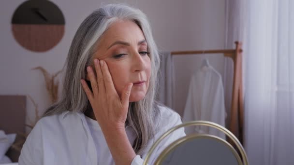 50s yeas stará dáma s šedými vlasy těší kůže vlhkost při pohledu na zrcadlo odraz dotek obličeje krém nanášení 60s věk senior zralá žena kontrola kosmetologie výsledky obličejová kosmetika dotýkání tváře - Záběry, video