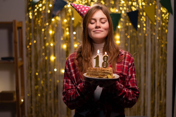Happy 18 years old girl in party cone make a wish and blowing out candles on birthday cake, celebrate birthday at home. Copy space - Foto, imagen
