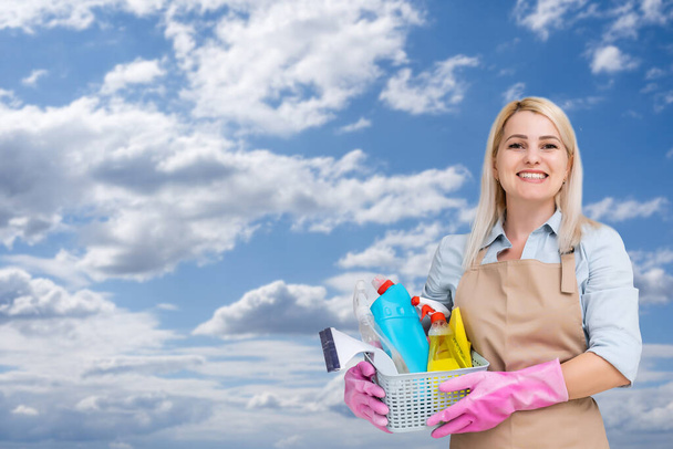 Молодая женщина держит чистящие средства и продукты в ведре, небо фон. - Фото, изображение