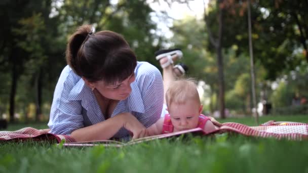 Äiti lukee kirjaa pikkutytölle makatessaan vihreällä ruoholla puistossa. 4K hidas Mo - Materiaali, video