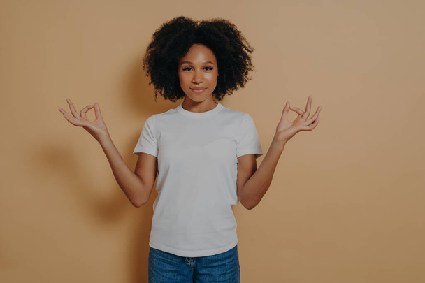 Koncept bdělosti. Mírumilovná afro-americká mladá žena v neformálním oblečení, která drží ruce v mudře a dívá se na fotoaparát s něžným úsměvem, je klidná a uvolněná, izolovaná na béžové stěně - Fotografie, Obrázek