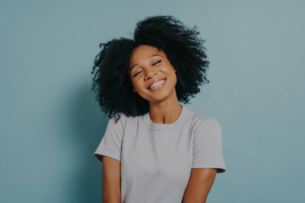 Beautiful overjoyed mixed race woman smiling and looking at camera. Portrait of cheerful young woman with afro hairstyle isolated over pastel blue studio background with copy space - Fotografie, Obrázek
