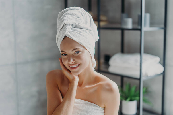 Beautiful European woman with makeup touches skin, has minimal makeup, has healthy glowing skin, wrapped in bath towel, enjoys rest at home. Spa woman poses in bathroom. Beauty, wellness concept - Photo, Image