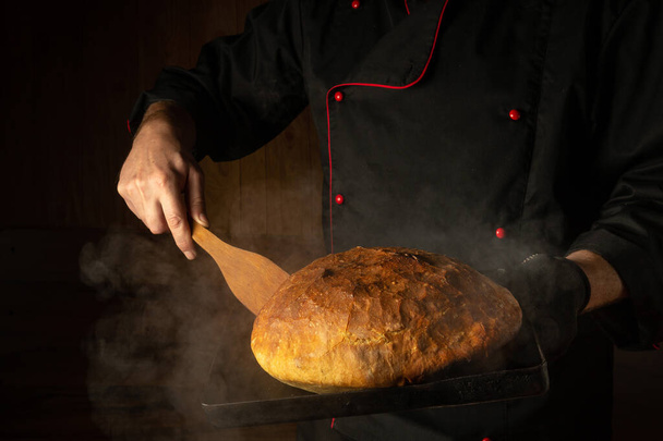 Der Bäcker backt in einer Bäckerei Brot auf einem Blech. Schaber in der Hand des Kochs. Platz für Menü oder Rezept auf dunklem Hintergrund - Foto, Bild