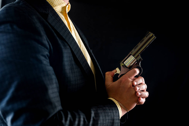 Ein Mann im Smoking und Hemd ohne Gesicht hält einen Revolver in den Händen vor der Brust, isoliert auf schwarzem Hintergrund. männliche Hände mit einer Waffe. Hände vor der Brust mit dem Revolver - Foto, Bild