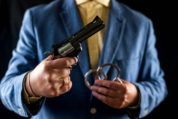 cropped portrait of male bodyguard in tuxedo holding weapon for defense. a bodyguard in a stylish tuxedo. revolver in hands. close up photo isolated on black background - Photo, Image