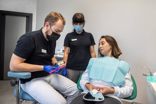 férfi fogorvos és egy női asszisztens megmutatják a betegnek, hogyan kell megfelelően megmosni a fogát és gondoskodni róluk. a fogorvos megmutatja az állkapcson, hogyan kell helyesen megmosni a fogat. fogászat egészséges fogak - Fotó, kép