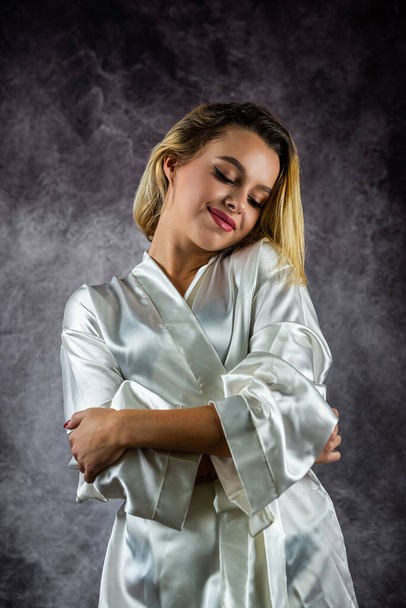Portret van een mooi meisje met mooi haar in een wit zijden gewaad glimlachend. zittende houding op studio achtergrond. geïsoleerd op een effen achtergrond - Foto, afbeelding