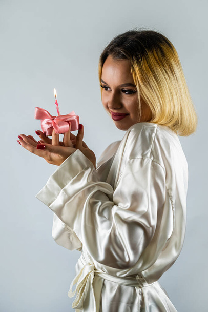 Модная девушка в шелковом белом халате задувает свечи на кусочке кекса на изолированном фоне. день рождения. праздник - Фото, изображение