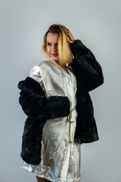 красивая молодая женщина в красивом легком шелковом халате с теплым меховым пальто сверху. изолированы на дымном фоне. концепция тепла и богатства. женский портрет - Фото, изображение