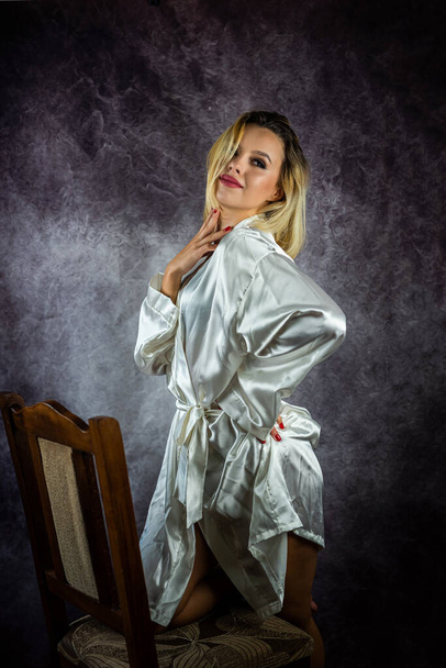 Красивая взрослая сексуальная женщина, сидящая на стуле в белом шелковом халате, позирующая в студии. изолированные на общем фоне. студия - Фото, изображение