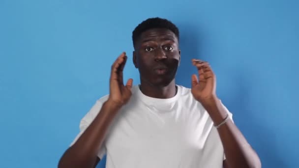 Afro-Amerikalı bir adam çığlıklarla boğuşuyor ağzı ardına kadar açık ellerini izole edilmiş mavi bir arka planda tutuyor. - Video, Çekim