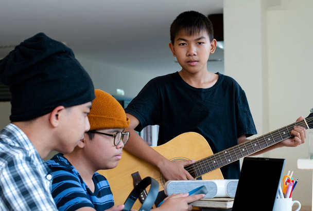Group of young asian teenagers sitting together inside the room working and practising school project about the music subject through laptop and playing quitar, too, soft and selective focus. - Foto, imagen