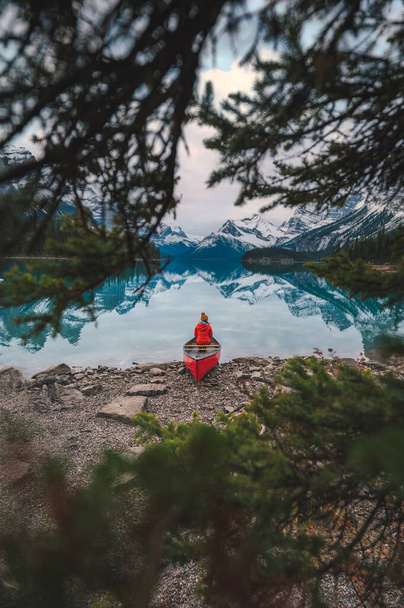 Joven viajero asiático en chaqueta roja sentado en canoa junto al lago en Spirit Island ubicado en el lago Maligne en el parque nacional Jasper, AB, Canadá - Foto, imagen