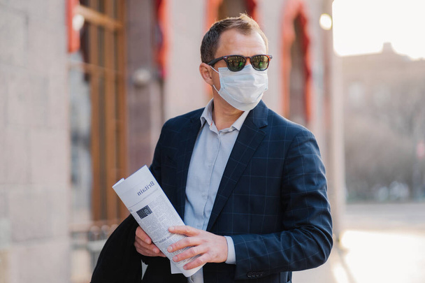 Businessman wears protective mask against transmissible infectious disease, reads newspapers, dressed elegantly, poses outdoor, thinks how to prevent new coronavirus from China. Flu in city. - Photo, Image