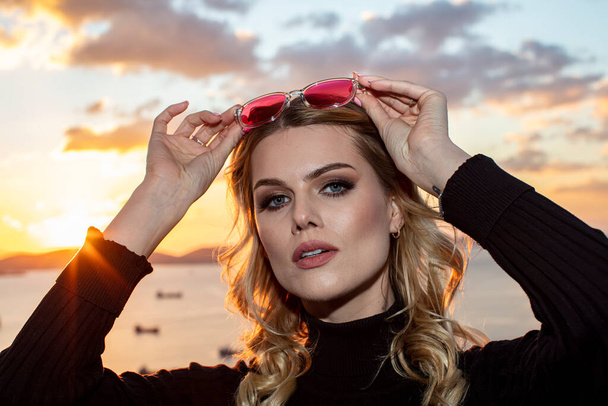 Beautiful female model wearing colorful glasses at sunset. Outdoors romantic portrait of attractive blonde woman with makeup and glasses posing. Istanbul archipelago (Princess Islands) skyline. - Photo, Image