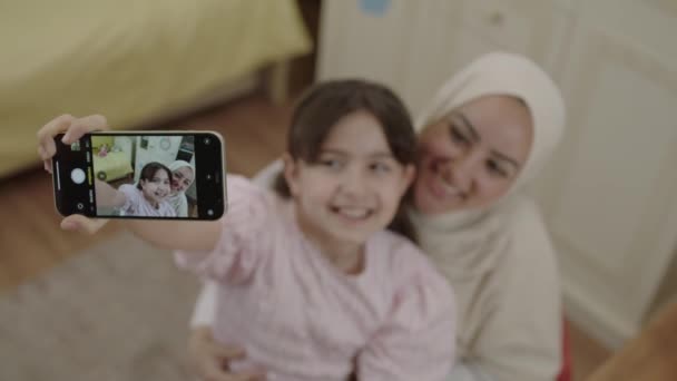 Маленькая милая девочка делает селфи со своей молодой матерью в хиджабе, записывает видео, фотографирует прекрасный момент. Мама смотрит в камеру, а дочь наслаждается видеозвонком. Вид фронтальной камеры телефона. - Кадры, видео