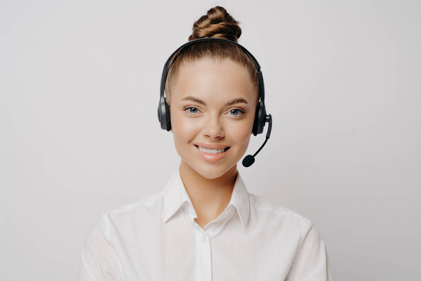 Hymyilevä asiantunteva nainen Call Center työntekijä valkoinen paita hiukset pulla ja musta kuulokkeet mielellään palvella ja auttaa asiakkaita, seisoo yksin edessä harmaa tausta - Valokuva, kuva