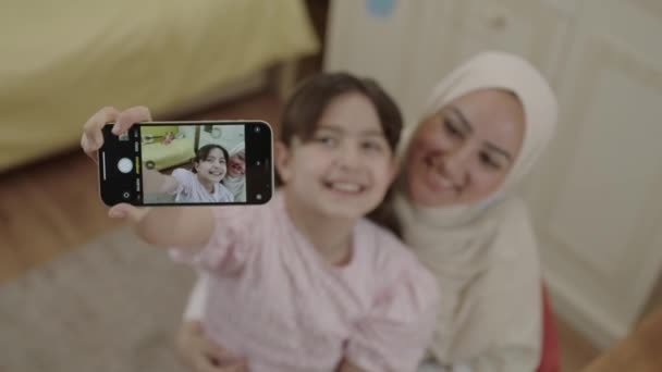Little cute girl taking selfie with her young mother in hijab, recording video, photographing beautiful moment. Mother looking at camera while daughter enjoying video call. Front phone camera view. - Video, Çekim