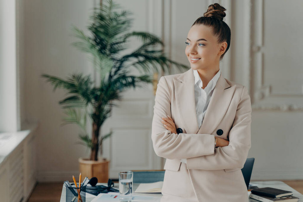 молодая веселая белая деловая женщина, одетая в формальный элегантный наряд, позирует у стола в светлом офисе и смотрит в сторону со счастливым выражением лица, чувствуя удовлетворение проделанной работой - Фото, изображение