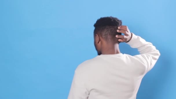 ein junger afroamerikanischer Mann wendet sich der Kamera zu, kratzt sich am Kopf und nickt vor blauem Hintergrund. - Filmmaterial, Video