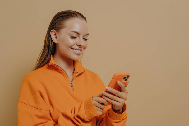 Happy charming young woman holding smartphone in hands and smiling, chatting in social networks, pleasant female in orange sweatshirt using mobile phone while standing isolated on beige background - Photo, image