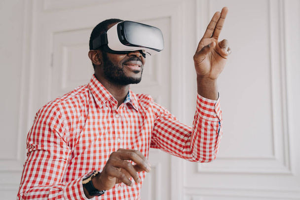 Enthusiastic smiling african ethnicity guy in VR headset glasses enjoying virtual reality online trying cyber world simulation at work, raised hands touching air with fingers, having fun with 3D gadget - Foto, Imagen