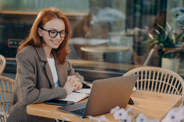 Redhead žena na volné noze hodinky trénink webinář dělá poznámky v notepadu sedí před otevřeným notebookem oblečený formálně pózuje ve venkovní kavárně prohlíží internet vyvíjí nový projekt - Fotografie, Obrázek