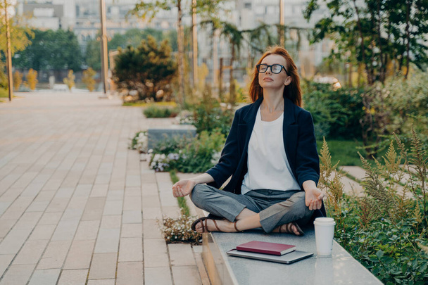 Спокійна ділова жінка, що медитує в лотосі на відкритому повітрі, мирний жіночий позаштатний працівник, що практикує вправи з йоги, сидячи в парку після важкого робочого дня в офісі, тримаючи руки в мудрах
 - Фото, зображення