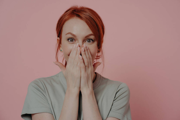 Young pretty ginger woman covering mouth with both hands, being in shock and disbelief, feeling amazed and cannot believe her eyes while posing isolated over pink background. Human emotions - Photo, Image