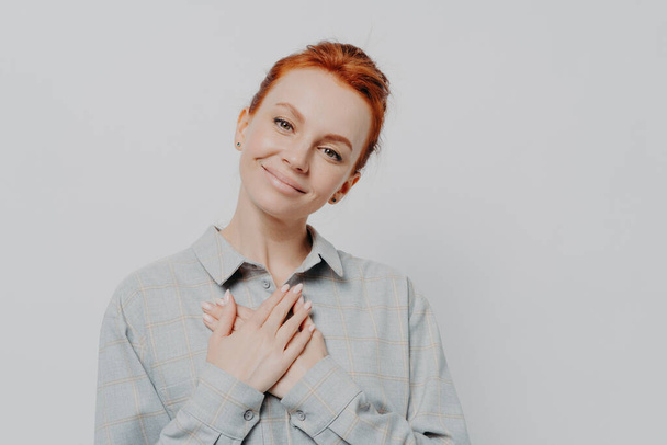 Young lovely red haired woman wearing shirt holding folded hands on chest and feeling grateful and thankful and support, looking at camera with charming smile, isolated on studio grey background - Photo, Image