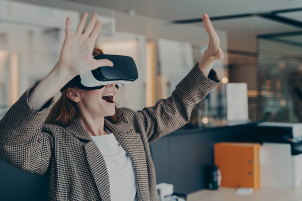 Indrukwekkende roodharige zakenvrouw gekleed in een draagbare VR-bril die de virtuele wereld verkent terwijl ze op haar werk zit en positieve emoties uitdrukt met gebaren - Foto, afbeelding