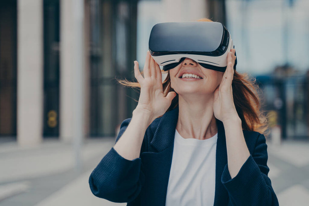 Удивительная рыжеволосая офисная работница, которая радуется, испытывая очки VR, тестируя виртуальную реальность для бизнеса, исследуя цифровой мир, стоя в одиночестве на фоне размытого городского фона - Фото, изображение