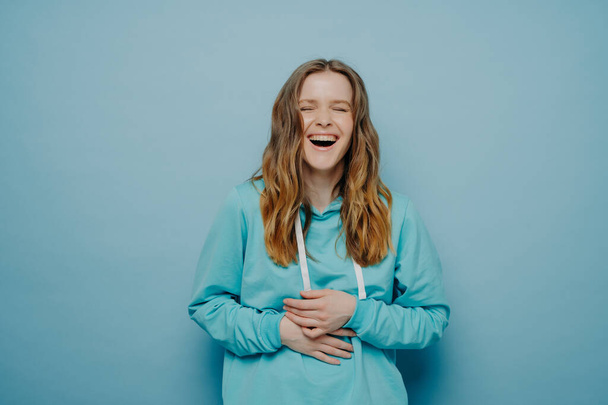 Overjoyed young woman dressed in hoodie holding belly and cant stop laughing after hearing hilarious joke, keeping eyes closed and having fun while standing isolated over blue background, studio shot - Photo, Image