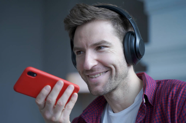 Close up of cheerful young german man in wireless headphones holding smartphone in hand with microphone close to his face during mobile conversation with friend, recording audio message - Photo, Image