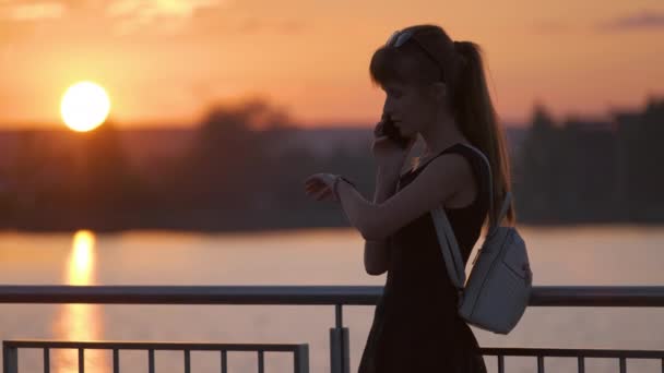 Jonge vrouw op de oever van het meer in het zomerpark praten op haar mobiele telefoon buiten op warme avond. Communicatie- en mobiele aansluitingsconcept. - Video