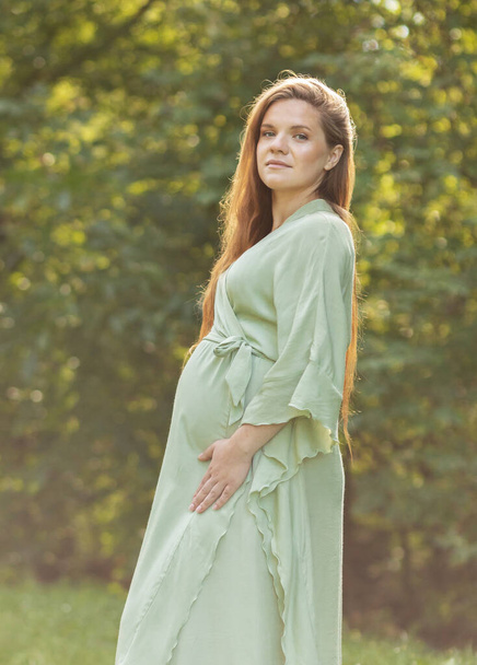 Young pregnant woman stands in park,looks at camera, green trees on background. Brown haired female wears dress.Magic happy pregnancy. Childbirth preparation, emotional connection with baby. Vertical - Foto, imagen