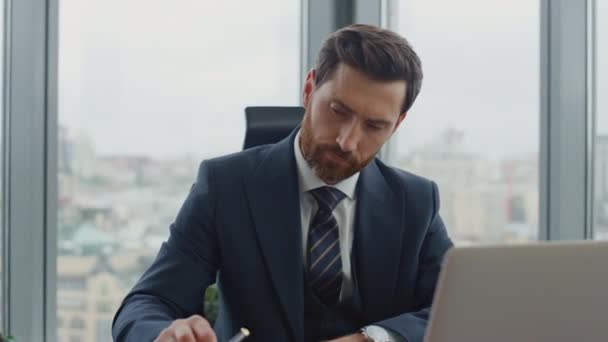 Thoughtful accountant writing financial papers sitting at work desk company office close up. Attractive bearded ceo manager worker thinking at corporate profit bank bills credits looking laptop screen - Footage, Video