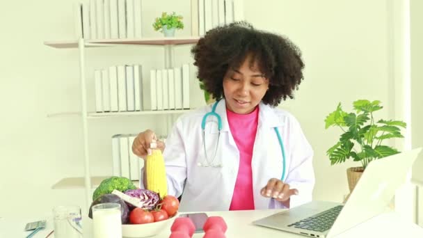 Schattige Afrikaanse Amerikaanse vrouwelijke voedingsdeskundige zit op haar bureau glimlachend en presenteren organische plantaardige maïs online aan de patiënt te consumeren op laptop computer werken in het ziekenhuis kantoor. - Video