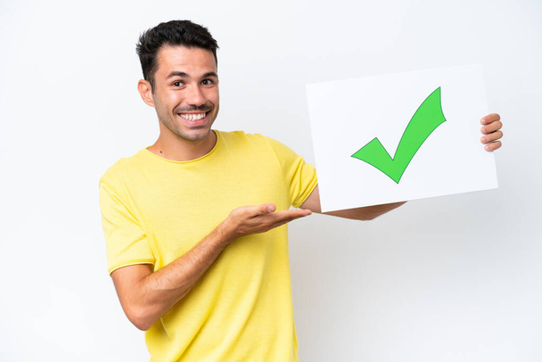 Young handsome man over isolated white background holding a placard with text Green check mark icon and  pointing it - Photo, Image