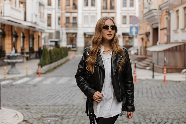 Modna blondynka model z czarną skórzaną kurtką i okularami przeciwsłonecznymi spacerując ulicą miasta - Zdjęcie, obraz