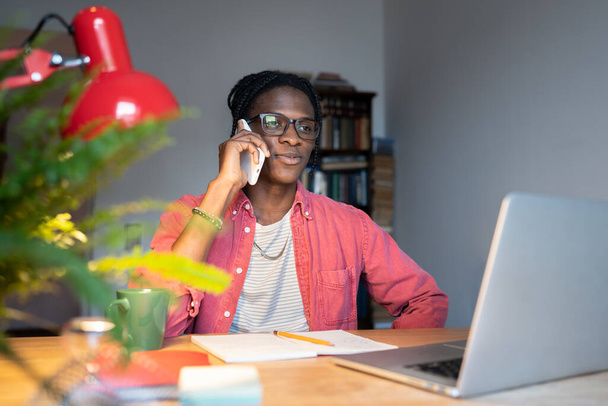 African guy wearing glasses talking on phone while sitting at desk with laptop, distracted from online study. Young black man holding smartphone speaking with friend while studying remotely at home - Photo, Image