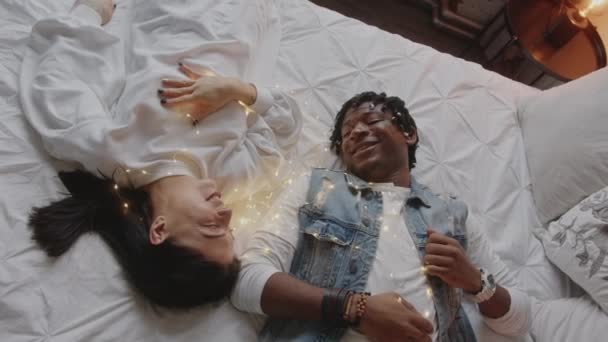 Ein multiethnisches Paar liegt im Bett, lacht und spielt mit der Weihnachtsbeleuchtung. Hochwertiges 4k Filmmaterial - Filmmaterial, Video