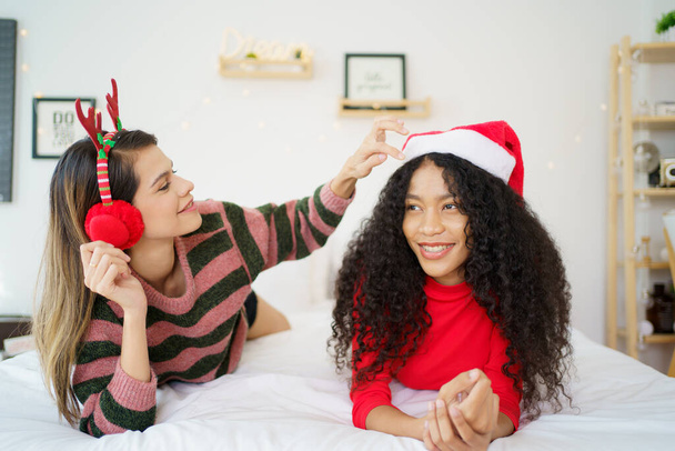 Heureuses femmes noires et caucasiennes gaies habillées dans le thème de Noël ou du Père Noël et allongées sur le lit. - Photo, image