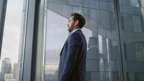 髭を生やした会社の従業員は、街の景色を眺めながらオフィスの窓の近くに立っているビジネスの視点を考えるストレスを感じています。金融危機の解決だけを心配する優雅な実業家 - 映像、動画