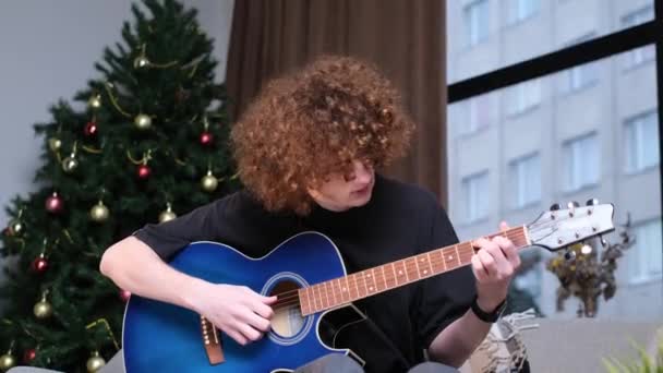 Ganzes Porträt eines lässigen Typen mit lockigem Haar, der vor dem Hintergrund des Weihnachtsbaums Gitarre spielt. - Filmmaterial, Video