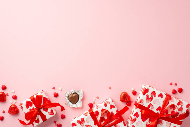 Концепция Дня Святого Валентина. Верхний вид фото подарочных коробок в оберточной бумаге с рисунком сердца шоколадные конфеты и посыпки на изолированном светло-розовом фоне с копирайтом - Фото, изображение