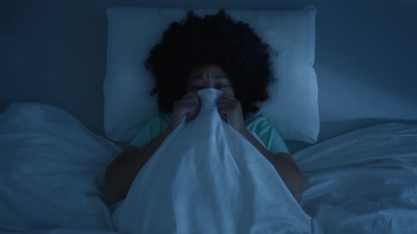 Visão superior sobre a mulher de cor deitada na cama e cobrindo rosto por cobertor para não ver coisas assustadoras em casa 4K. Assustada afro-americana com medo de fantasmas na cama, escondida debaixo do cobertor na noite escura - Filmagem, Vídeo